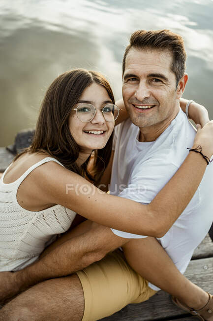 Sorrindo filha abraçando pai enquanto sentado no molhe — Fotografia de Stock
