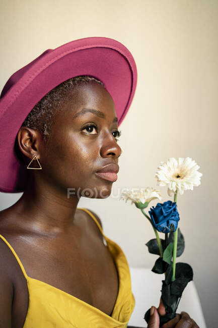 Hipster femelle réfléchie portant chapeau rose tenant des fleurs tout en regardant loin à la maison — Photo de stock