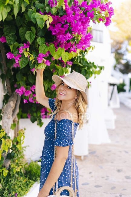 Smiling female tourist touching flowers growing on plant in Binibeca village, Minorca, Spain - foto de stock