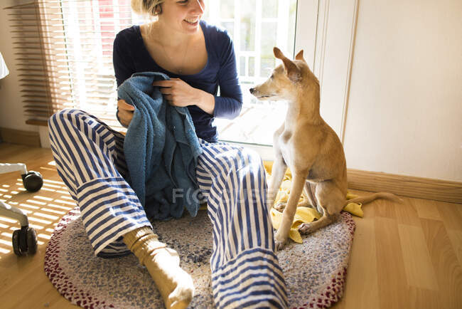 Усміхнена жінка з рушником навчає собаку, сидячи біля вікна вдома. — стокове фото