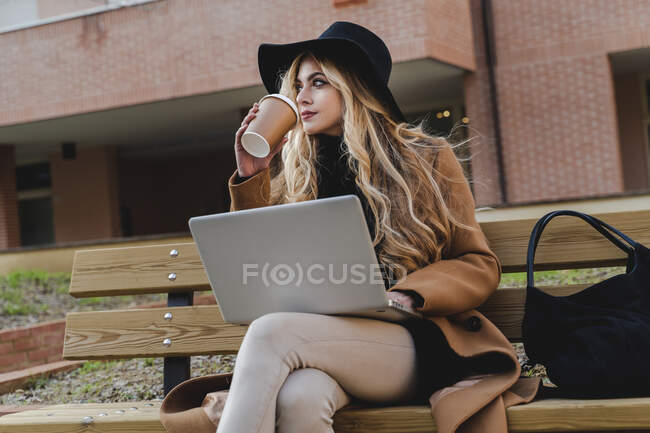 Donna con computer portatile che beve caffè guardando altrove — Foto stock