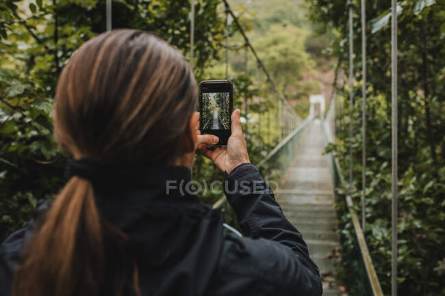 Woman photographing with mobile phone on suspension bridge in forest — Stock Photo