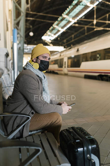 Uomo con maschera viso e telefono cellulare in attesa mentre seduto sulla panchina alla stazione — Foto stock