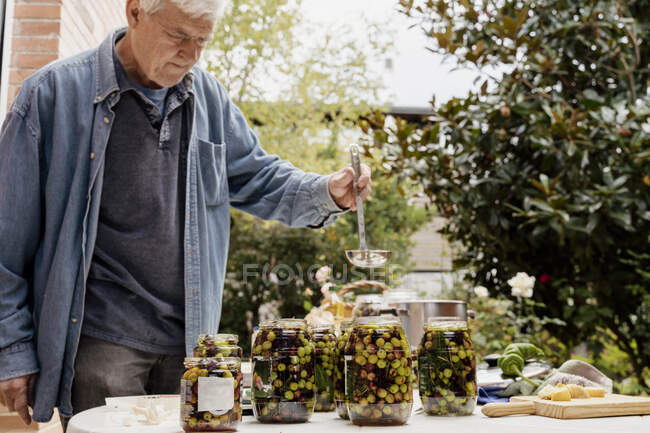 Hombre mayor preparando aceitunas en frascos en la mesa en el patio trasero - foto de stock