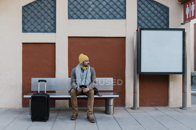 Mid adult man wearing knit hat using digital tablet while sitting on bench at station — Stock Photo