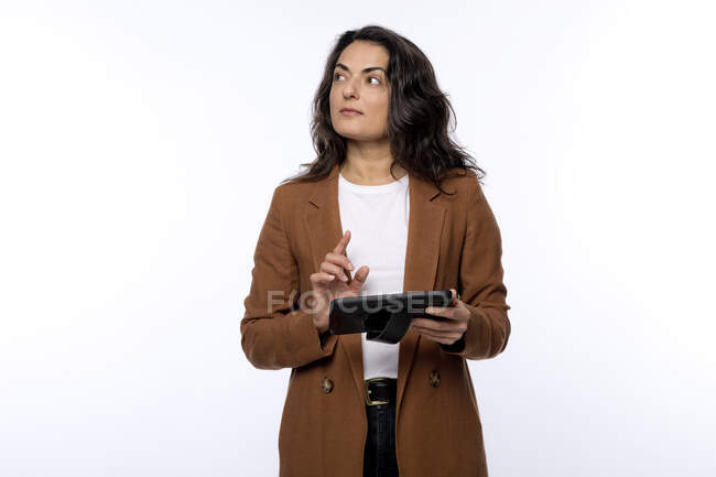 Mujer madura reflexiva con tableta digital mirando hacia otro lado sobre fondo blanco - foto de stock