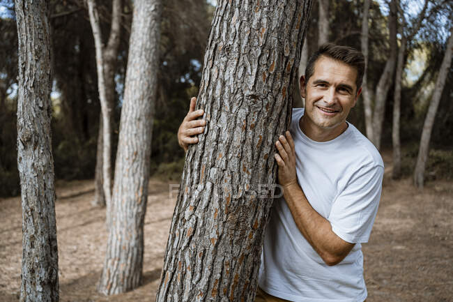 Hombre sonriente abrazando tronco de árbol en el bosque durante las vacaciones - foto de stock