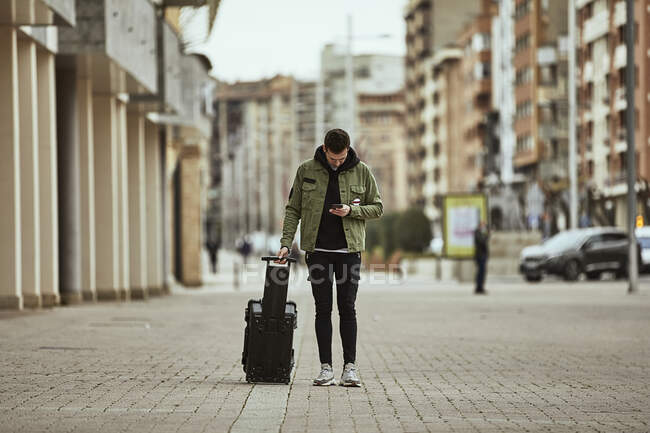 Young man with suitcase looking at mobile phone while standing on footpath in city — Stock Photo