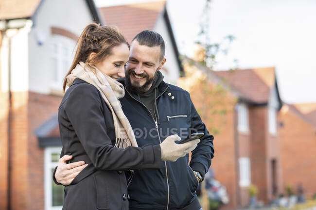 Hombre sonriente mirando a su novia usando el teléfono inteligente mientras está de pie contra nuevas casas - foto de stock