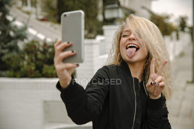 Jovem feliz tomando selfie enquanto a paz gesto e furar a língua através do telefone inteligente — Fotografia de Stock