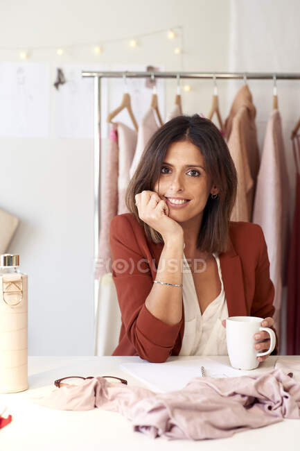 Smiling female fashion entrepreneur sitting with hand on chin at desk in design studio — Stock Photo