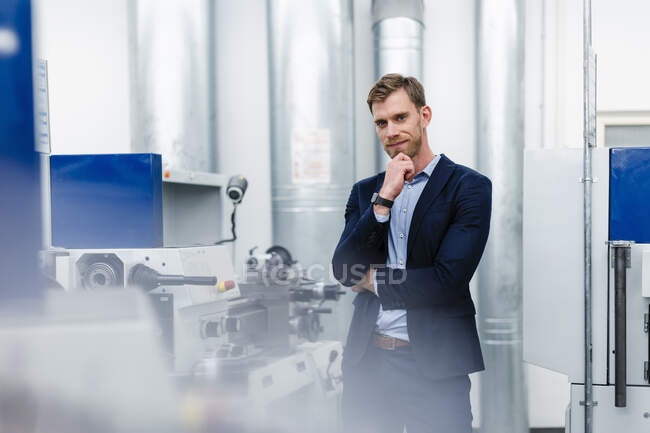 Smiling businessman with hand on chin standing by machinery equipment in factory — Stock Photo