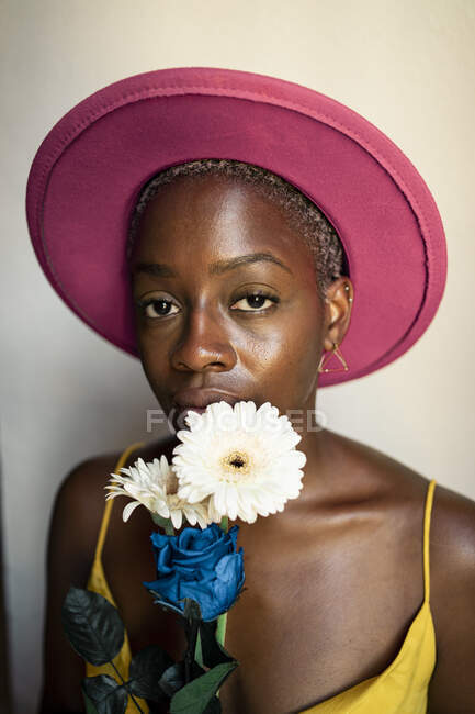 Confiada hipster femenina con sombrero rosa sosteniendo flores contra la  pared en casa — Etnia afroamericana, Azul - Stock Photo | #481862838