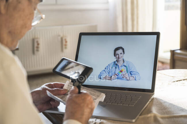 Senior man reading through magnifying glass while female doctor consulting on video call — Stock Photo