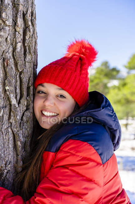 Young woman wearing red knit hat smiling while hugging tree standing in forest — Stock Photo