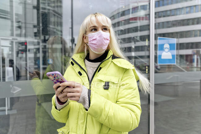 Jeune femme portant un masque facial à l'aide d'un téléphone intelligent tout en se tenant contre le mur de verre — Photo de stock