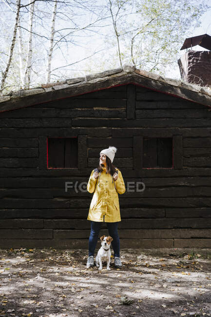 Frau im gelben Regenmantel steht mit Hund vor Blockhaus — Stockfoto