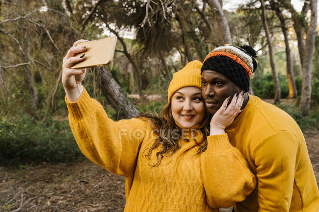 Giovane donna che indossa cappello maglia prendendo selfie attraverso il telefono cellulare mentre in piedi alla foresta — Foto stock