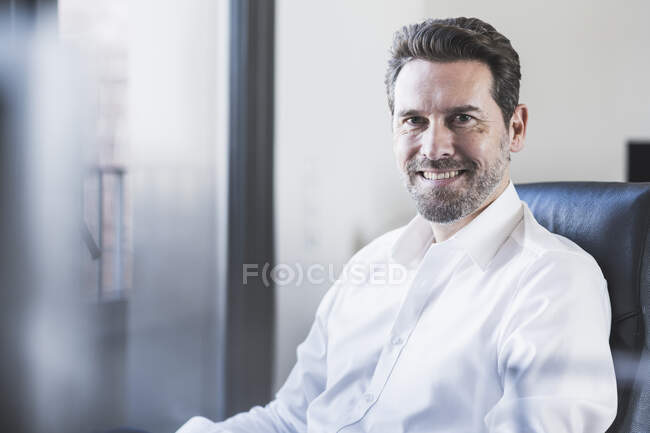 Hombre de negocios confiado sonriendo mientras está sentado en la silla en la oficina - foto de stock