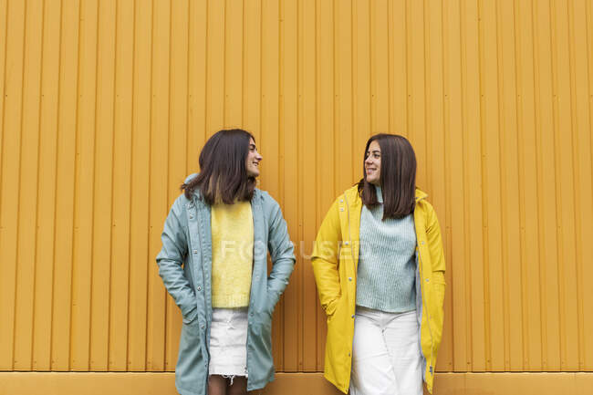 Sisters wearing yellow and blue raincoats smiling while looking at each other standing against wall — Stock Photo
