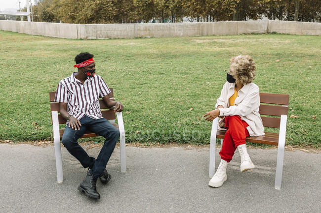 Freunde mit Mundschutz reden, während sie mit sozialer Distanz auf einem Stuhl sitzen — Stockfoto