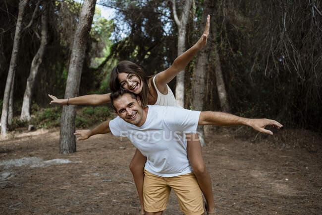 Sonriente padre dando paseo a cuestas a su hija con los brazos extendidos en el bosque durante las vacaciones - foto de stock