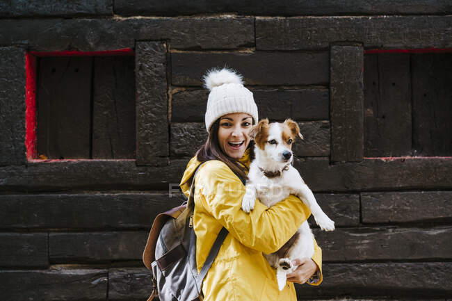 Портрет усміхненої жінки в жовтому плащі, що тримає собаку перед зрубом — стокове фото