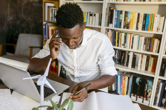 Чоловічий еколог з моделлю вітрових турбін розмовляє на смартфоні в домашньому офісі — стокове фото