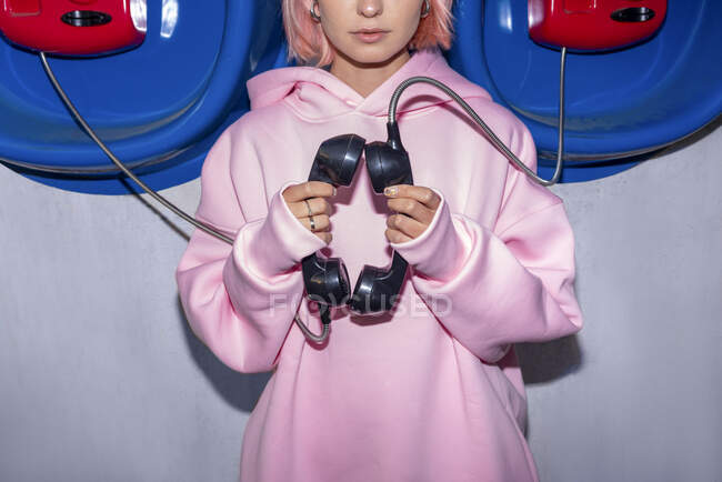 Young woman wearing pink hooded shirt standing in front of telephone booths, holding receivers — Stock Photo