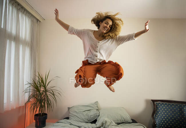 Allegro giovane donna con le braccia alzate saltando sopra il letto in camera da letto — Foto stock