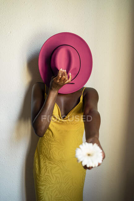 Hipster femenino sosteniendo flor blanca mientras cubre la cara con sombrero rosa contra la pared en casa - foto de stock