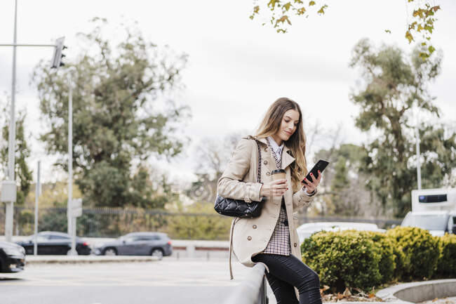 Donna alla moda con tazza di caffè usa e getta utilizzando smart phone mentre seduto su ringhiera in strada — Foto stock