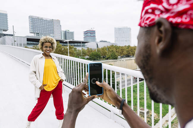 Чоловік фотографує подругу через мобільний телефон стоячи з руками в кишенях на мості в місті. — стокове фото