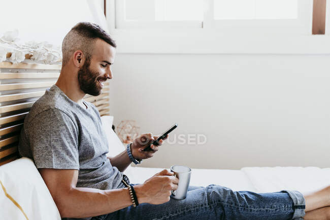 Giovane sorridente che lavora sul cellulare seduto sul letto a casa — Foto stock