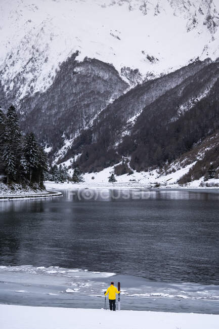 Joven con esquí en la orilla del lago cubierta de nieve en frente de las montañas - foto de stock