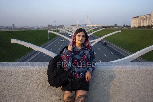 Hipster female with back pack leaning on retaining wall at bridge — Stock Photo