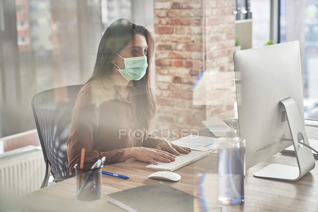 Femme adulte moyenne travaillant sur ordinateur sur bureau vu à travers le verre pendant la pandémie — Photo de stock