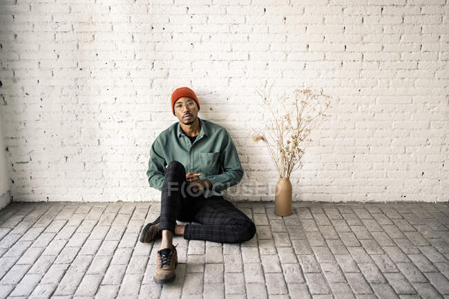 Mittlerer erwachsener Mann mit leerem Gesichtsausdruck sitzt an getrockneter Pflanzenvase vor weißer Ziegelwand — Stockfoto