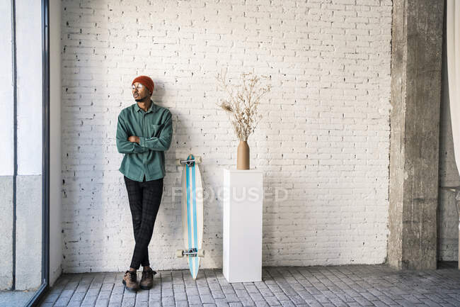 Homem pensativo com braços cruzados de pé por skate contra parede de tijolo branco — Fotografia de Stock