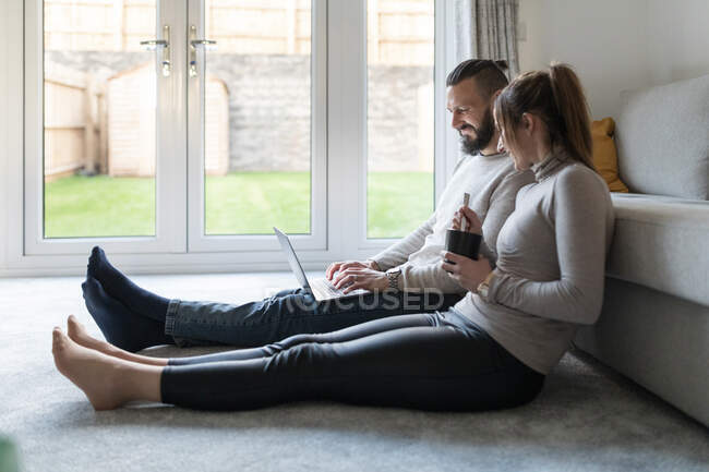 Sorrindo jovem mulher tomando chá enquanto namorado usando laptop em casa — Fotografia de Stock