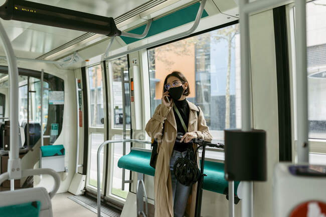 Femme avec scooter électrique de poussée et masque parlant sur le téléphone portable tout en se tenant dans le tram — Photo de stock