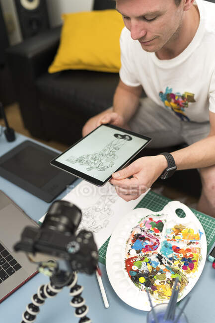 Artista mirando la tableta digital mientras transmite en vivo a través de un ordenador portátil sentado en casa - foto de stock