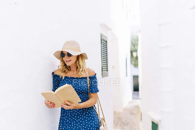 Mulher usando livro de leitura de chapéu enquanto estava em casa na aldeia de Binibeca, Minorca, Espanha — Fotografia de Stock
