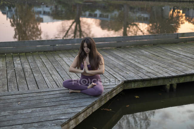Femme avec les cheveux longs méditant tout en étant assis sur la jetée au-dessus du lac au coucher du soleil — Photo de stock