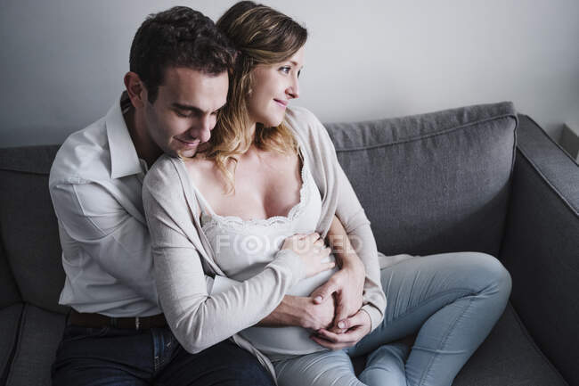 Мужчина с закрытыми глазами обнимает беременную жену, сидя дома на диване — стоковое фото