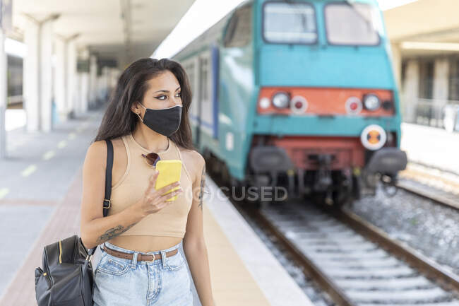 Jovem mulher usando máscara facial olhando para longe ao usar o telefone móvel em pé na estação — Fotografia de Stock