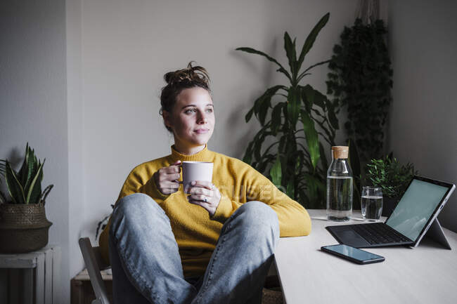 Mujer joven bebiendo café mientras está sentada junto a la mesa en casa oficina - foto de stock