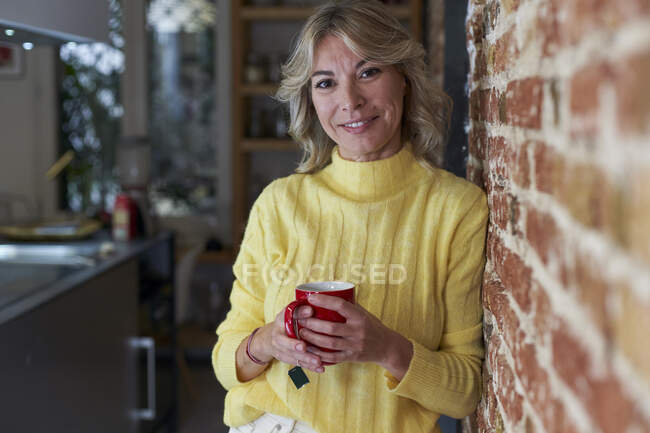 Sorrindo mulher madura inclinando-se na parede de tijolo enquanto segurando xícara de chá na cozinha — Fotografia de Stock