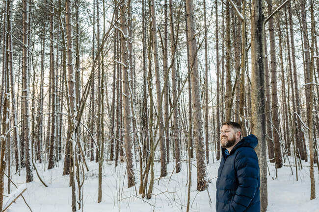 Усміхнений чоловік у теплому одязі, який відводить погляд у лісі. — стокове фото