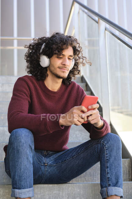 Giovane bell'uomo con le cuffie utilizzando smartphone mentre seduto su gradini — Foto stock